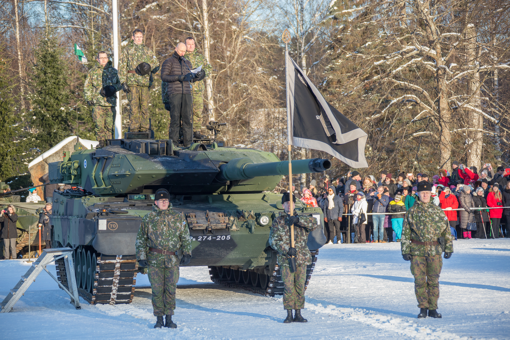 Sotilaat seisovat panssarivaunun päällä ja vierellä asennossa, yhdellä lippu kädessä, taustalla yleisöä.