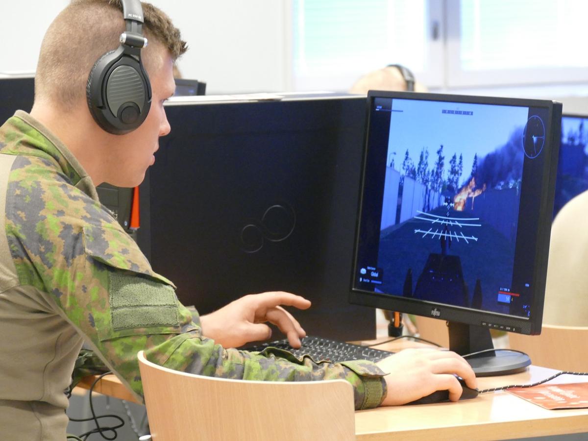 Upseerioppilas näytön edessä, hän käyttää tietokonepeliä muistuttavaa ohjelmaa sotilaskoulutuksessa. Näytöllä näkyy osuma ja räjähdys.